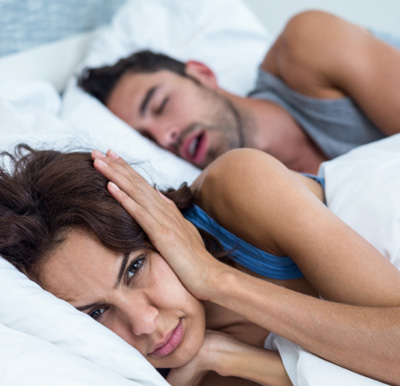 a woman covering her ears in bed with a man snoring behind her