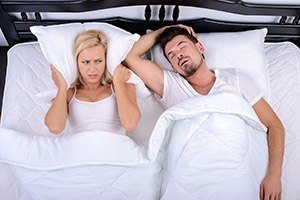 Frustrated woman covering ears next to snoring man in bed