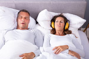 Man snoring in bed, woman covering ears with headphones